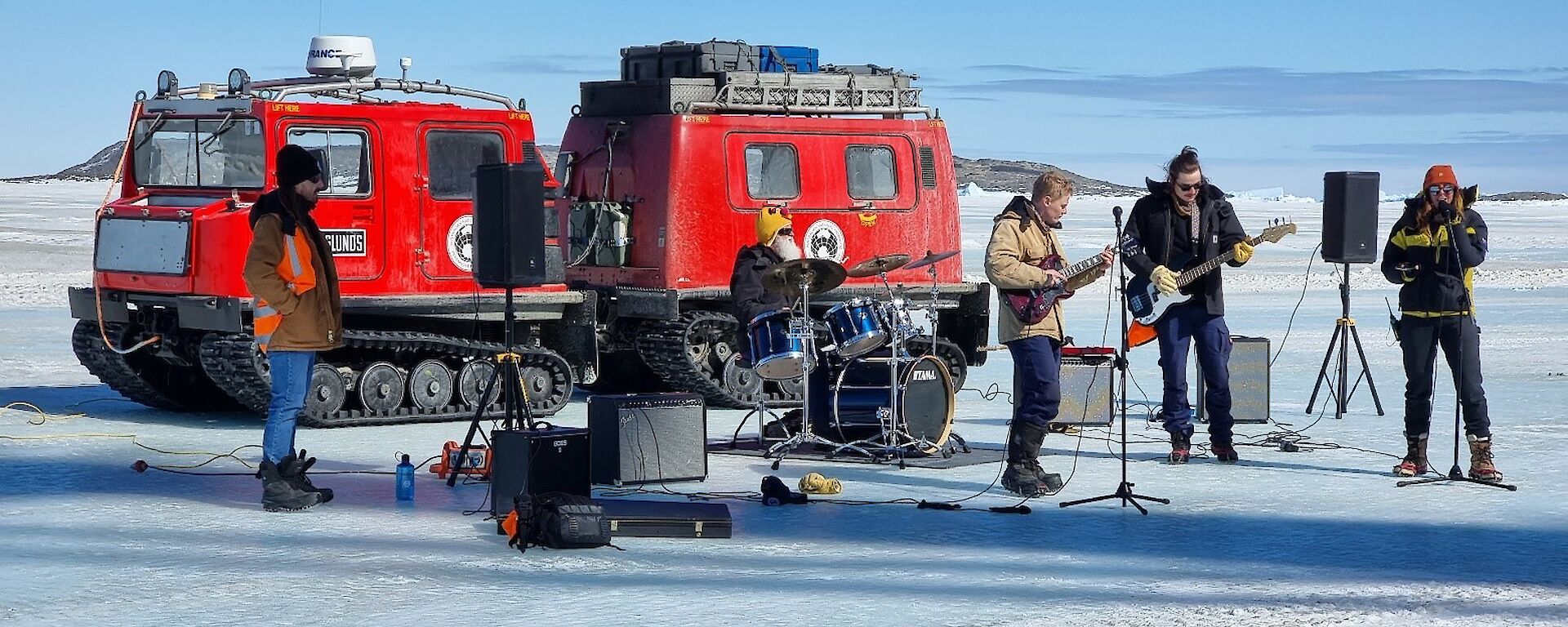 A group of musicians play, with a red snow tractor behind them on the ice