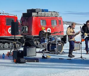 A group of musicians play, with a red snow tractor behind them on the ice