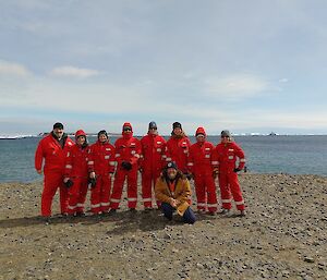 A group in red jumpsuits stands on a rocky shore with sea behind them