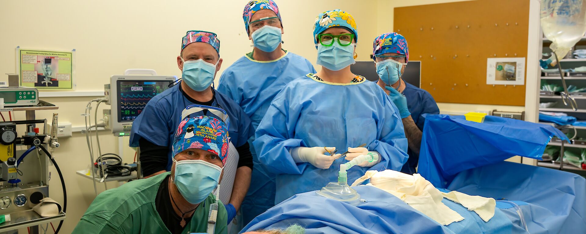 In a medical facility, five people are wearing medical PPE standing behind a person acting as the patient on the operating table.