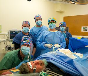 In a medical facility, five people are wearing medical PPE standing behind a person acting as the patient on the operating table.