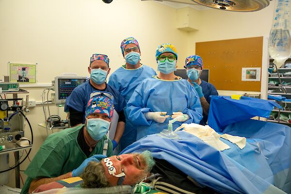 In a medical facility, five people are wearing medical PPE standing behind a person acting as the patient on the operating table.
