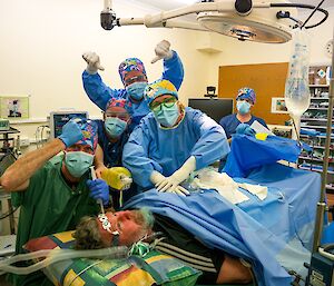 In a medical facility, five people are wearing medical PPE standing behind a person acting as the patient on the operating table.  They are pretending to resuscitate the patient.