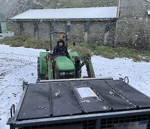 A man is driving a green tractor with a cage pallet on the front. It is snowing.