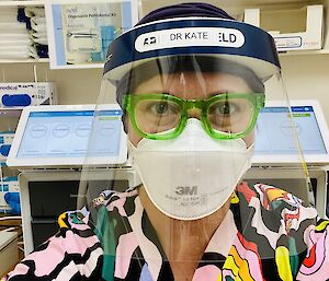 A selfie of a woman with green glasses and bright colourful scrubs. She has on a facemask and face shield.