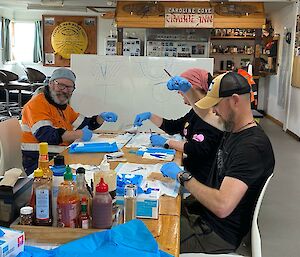 Three people sit around a table practicing their suturing. There is a whiteboard displaying the types of cuts which need to be fixed.