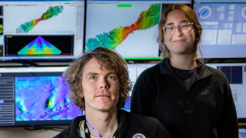 Two people sitting beside computer monitors with colourful seafloor maps on the screens.