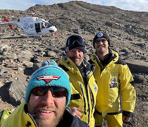 Three men in yellow stand in front of a hut on a rocky hill with a helicopter in the background