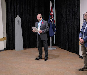 A man in a grey suit, with a red poppy on his lapel, stands in front of an Australian flag.
