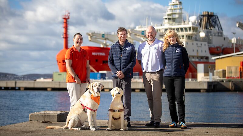 A group of people stand smiling in front of the icebreaker RSV Nuyina, with Murphy the guide dog and a plastic collection dog.