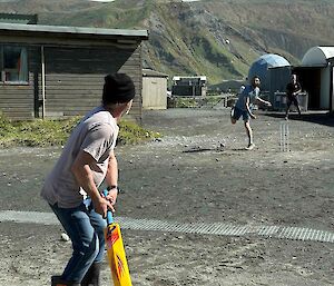 A person in gumboots stands with a cricket bat while another person bowls at them.