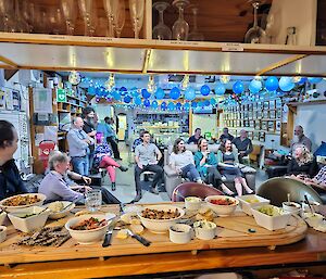 A table in the foreground with food on it, people sitting in chairs in the background.