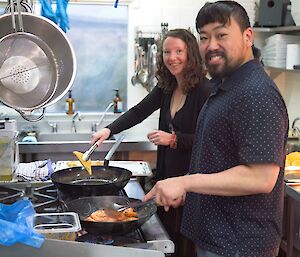 A man and a woman cooking with frypans in the kitchen.