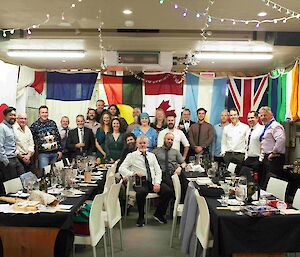 People stand around behind two long formal dining tables, with lots of flags behind them.