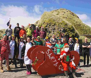 A group of people stand with a red Santa sled, some wearing penguin suits.