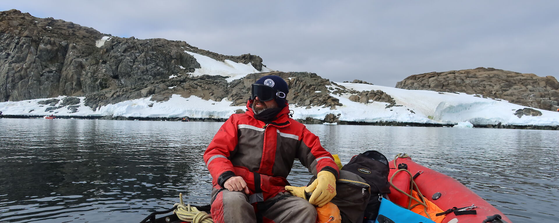 A man in red survival gear sits in a red rubber dingy on a still bay with snow and rocks behind him