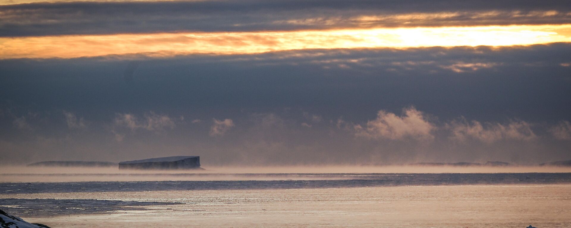 Sea smoke and associated fractostratus cloud west of the Bailey Peninsula, icebergs in background with colors associated with the setting of the sun piercing through clouds