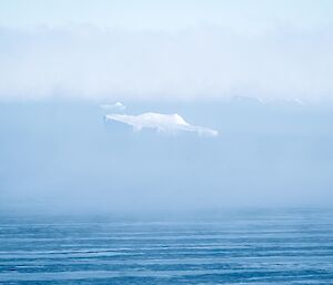 calm icy ocean with a backdrop of fog and a distant iceberg surfaces afar.
