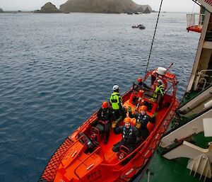 Five people in a small orange boat being lowered by crane into the sea from a larger ship.