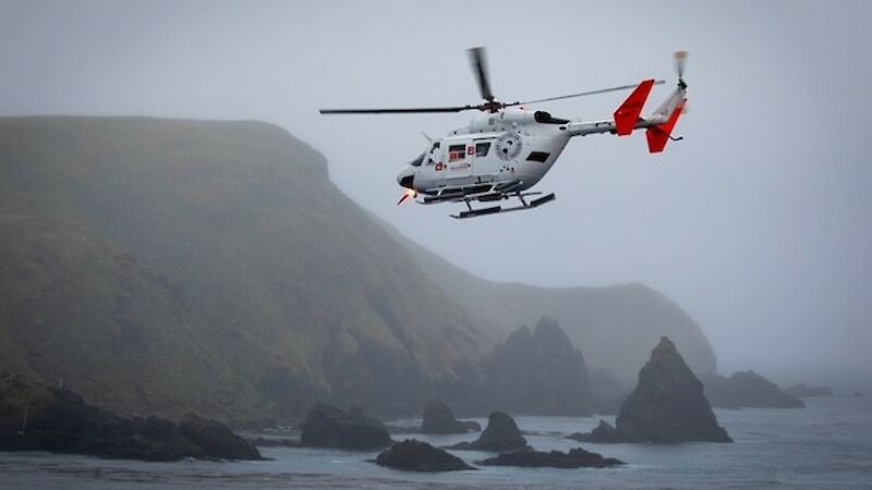 A helicopter in the air with Macquarie Island’s rugged hills behind it.