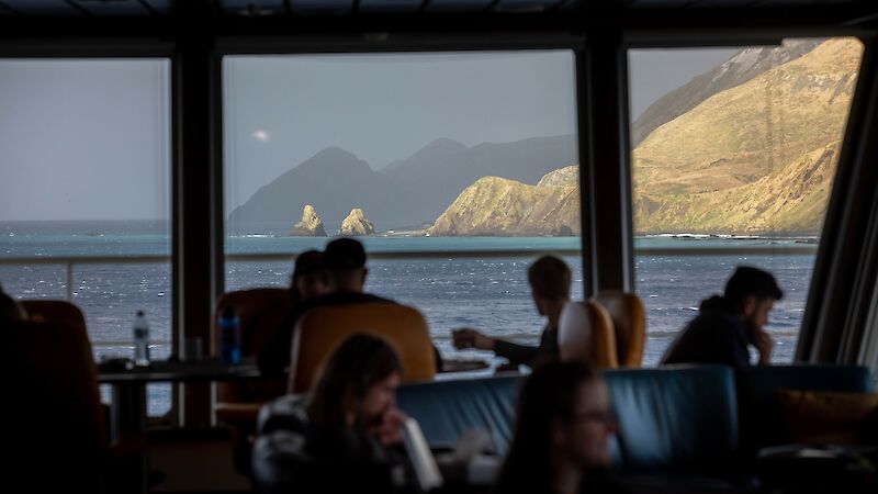 People sit in a ship’s lounge looking out the window to steep hills beyond
