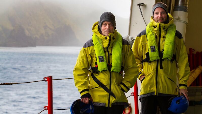 Two men in extreme weather gear stand on the deck of a ship with an island in the background.