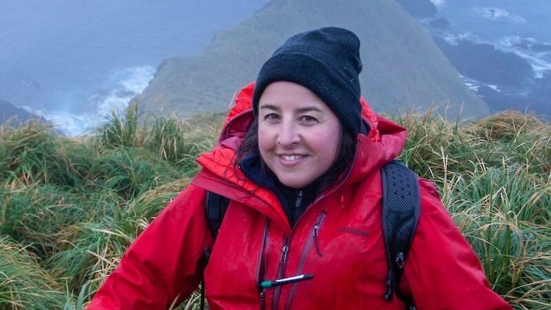 A smiling woman on a steep hillside wearing a red coat and black beanie.