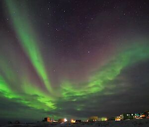 A green and purple aurora appears over a cluster of buildings in the darkness.