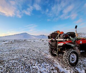 A quad bike parked on snow-covered pebbly ground with snow-covered hills in the distance.
