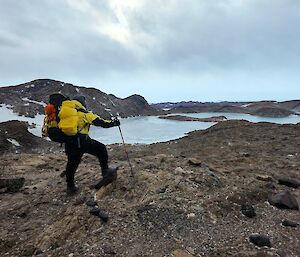 A man posed with his backpack of equipment on rocky ground beside an ice-covered lake.