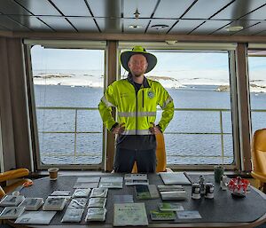 Expeditioner postmaster Owen stands behind table with post supplies aboard RSV Nuyina