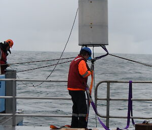 A man on the rear deck of a ship hauling in a large cylindrical scientific instrument that is hanging at head height beside him.