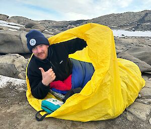 A man inside a yellow bivvy bag, lying on some rocks in Antarctica