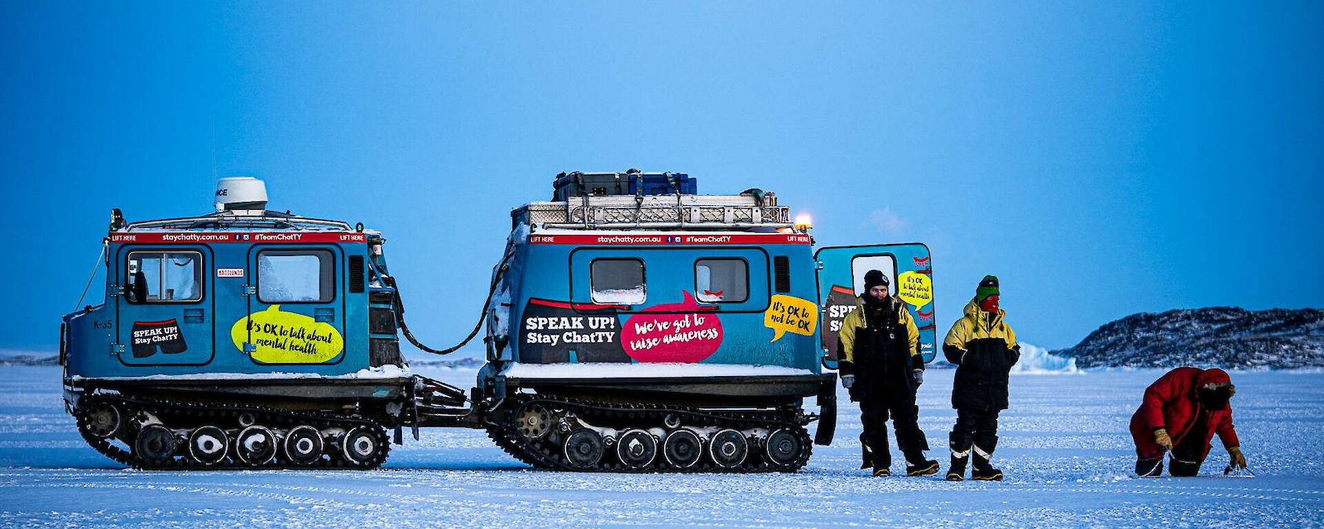 Three people stand beside a blue hagglund vehicle on snow.