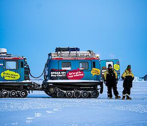 Three people stand beside a blue hagglund vehicle on snow.