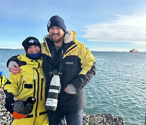 A man wearing a large camera around his neck, standing with his mother beside the ocean, with a ship in the background.