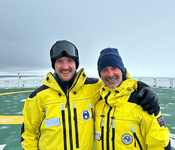 Two men in yellow jackets and black beanies on the helideck of a ship, with sea ice in the background.