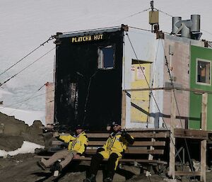 Two men sitting on a bench in front of a multi-coloured rectangular hut made from a shipping container.