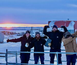 Four expeditioners pose on a metal walkway with a sunset over snow and ice behind.