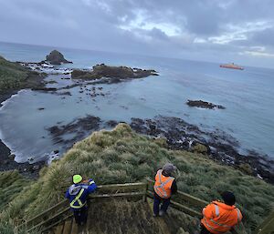Three expeditioners on platform look across water to the ship sailing north