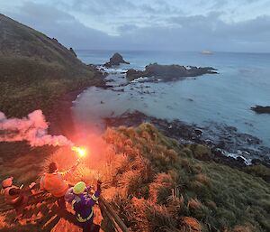 A group of expeditioners on a platform on a hill slope, look across water to the ship sailing north and one lets of a red flare