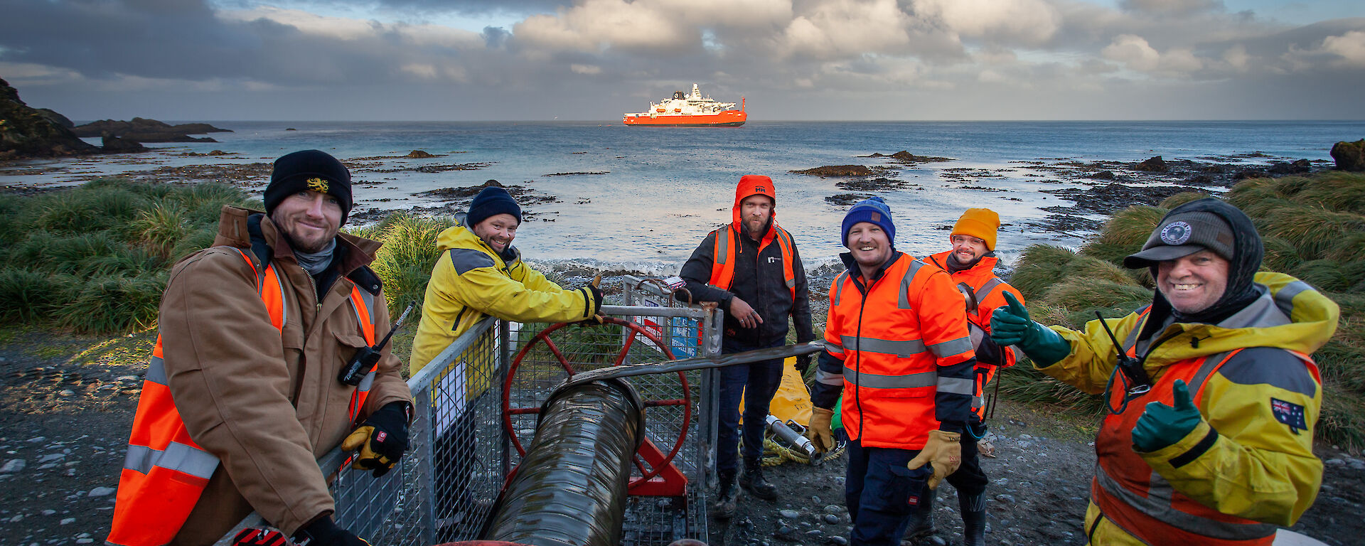 Men in high vis stand around fuel hose reel with water behind and ship on the horizen