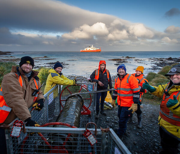 Men in high vis stand around fuel hose reel with water behind and ship on the horizen