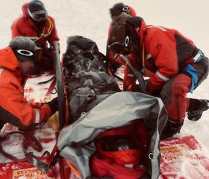 three team members wrapping the 'patient' up for extended sea ice retrieval using a stretcher, sleeping bag and tarp.