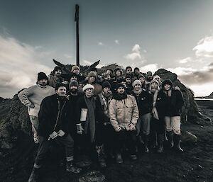 24 expeditioners standing and posing without smiles to camera, under the base of a large rock with anchor on top