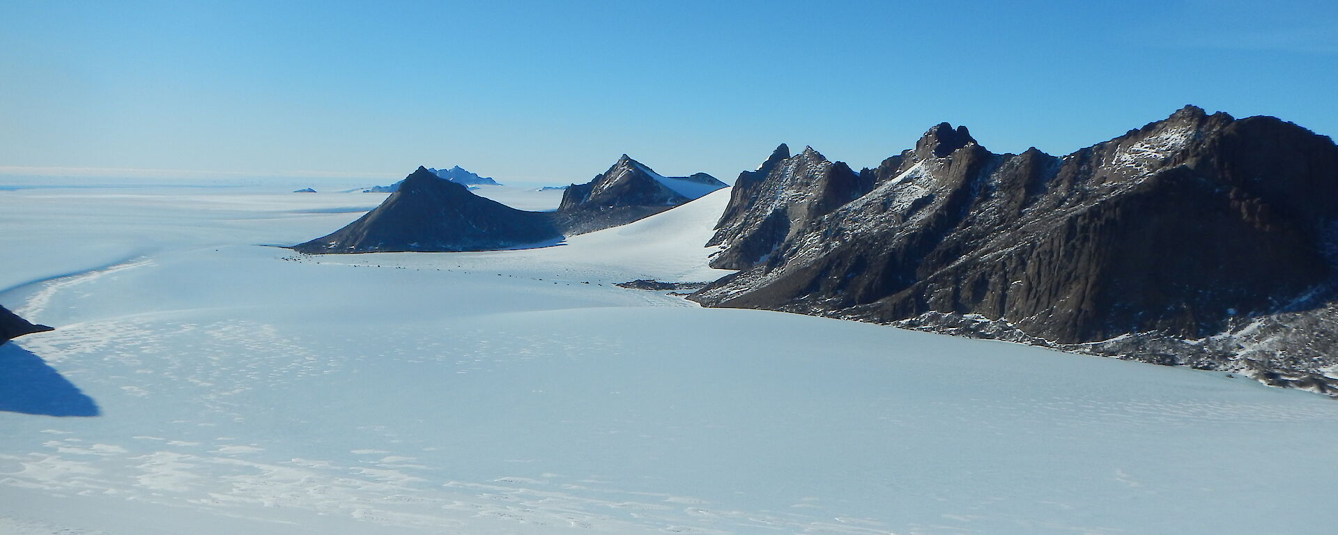A snowy flat plain with grey jagged mountains in the background