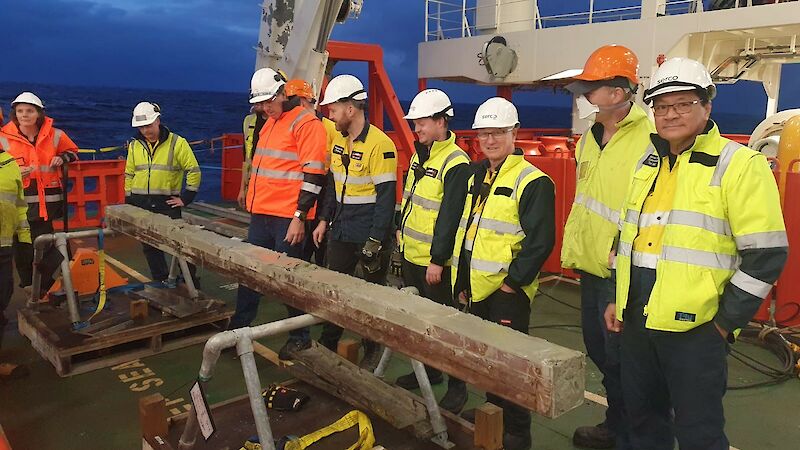 Eight people stand alongside a rectangular metal corer containing a four-metre long core of ocean sediment.