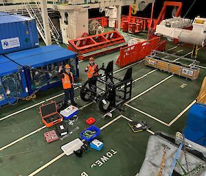 Two men in high visibility vests on the trawl deck of a ship, surrounded by tool boxes, preparing a metal cage containing a camera system for deployment