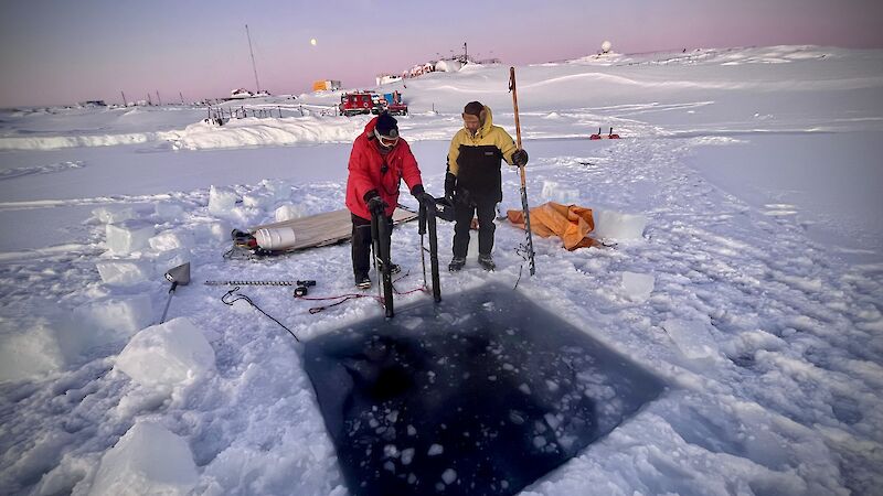 Two people look at a square hole in the ice with dark blue water in it.