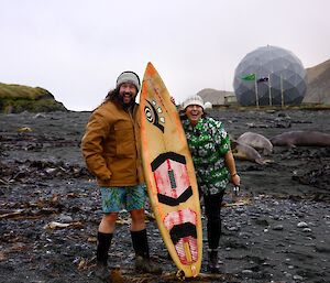 Two people, one in board shorts and jacket and one in a hawaiian shirt, hold the surf board and smile to camera